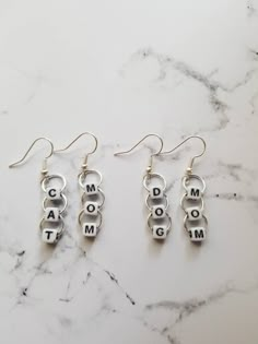 three silver earrings with the word mom and two charms hanging from them on a marble surface