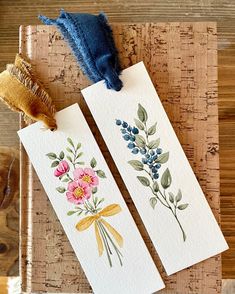 two tags with watercolor flowers on them sitting on a wooden table next to a tassel