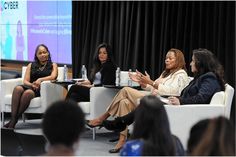 four women sitting on chairs in front of a screen talking to each other at an event