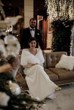 a man and woman in formal wear sitting on a couch next to each other with christmas decorations behind them