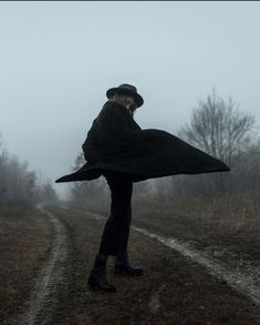 a person standing on a dirt road in the fog wearing a black coat and hat