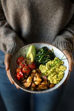 a person holding a bowl full of food and vegetables with avocado on top