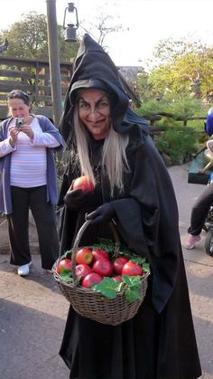 a woman dressed as a witch holding a basket of apples and vegetables in her hands