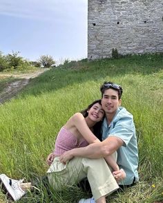 a man and woman sitting on the ground in front of an old castle with green grass