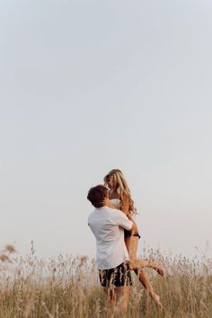 a man holding a woman in a field with tall grass and blue sky behind him