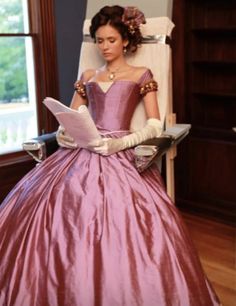 a woman in a pink dress sitting on a white chair reading a book while wearing gloves