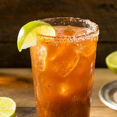 a tall glass filled with ice and lime on top of a wooden table next to sliced limes