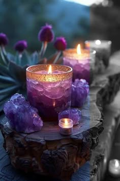 three candles are sitting on a log with purple rocks and flowers in the back ground