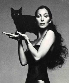 a woman holding a black cat in her right hand and looking at the camera with an angry look on her face