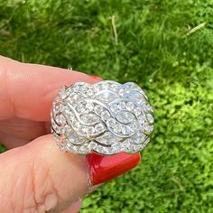 a hand holding a diamond ring in front of some green grass and bushes, with the top part of it being held up by someone's finger