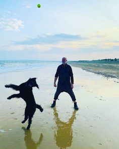 a man standing on top of a sandy beach next to a black dog jumping in the air