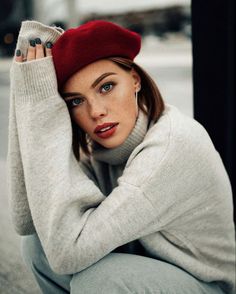 a woman in grey sweater and red beret sitting on the ground with her hands behind her head