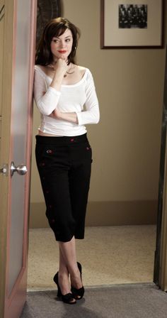 a woman in white shirt and black pants leaning against door with her hand on her chin