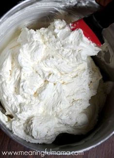 a metal bowl filled with whipped cream on top of a wooden table next to a red spatula