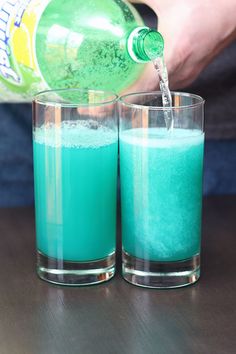 two glasses filled with blue liquid being poured into one glass and another is on the other side