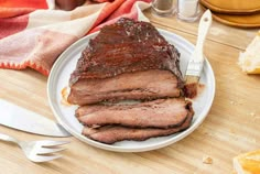 a white plate topped with sliced up meat next to bread and utensils on top of a wooden table