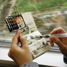 a person is holding an open book and some paintbrushes in their hand while looking out the window