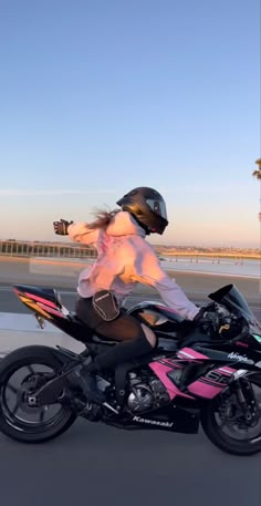 a woman riding on the back of a pink and black motorcycle down a street with palm trees in the background