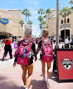 two women with pink backpacks walking down the street in front of a building and palm trees