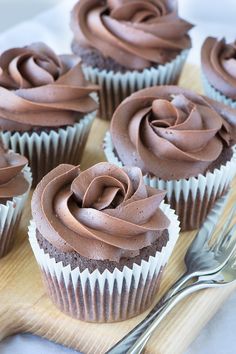 cupcakes with chocolate frosting on a wooden board next to a knife and fork