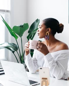 a woman sitting at a desk with a laptop and cup in her hand while holding a mug