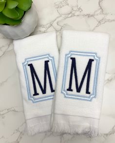 two white towels with the letter m on them sitting next to a plant and vase