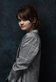 a woman standing in front of a black wall wearing a jacket and green earrings