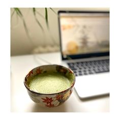 a bowl filled with green liquid sitting next to a laptop on a white table top