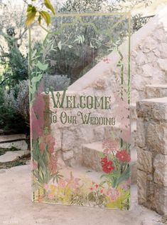 a welcome sign with flowers on it in front of a stone wall