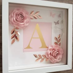 a white frame with pink paper flowers and gold foil leaves on it, in front of a wooden wall