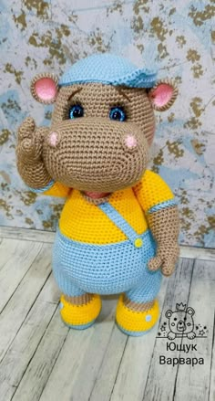 a crocheted hippo is standing on a wooden floor with a blue hat and yellow overalls