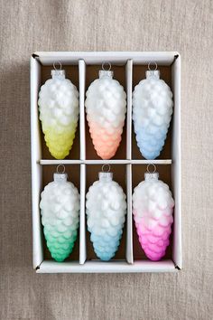 four different colored pine cone ornaments in a white box on a beige surface, one is hanging from the ceiling