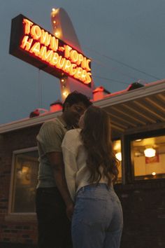 a man and woman standing in front of a town top hamburger restaurant at night with the neon sign lit up