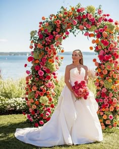 a woman in a wedding dress standing under an archway with flowers on the grass and water behind her