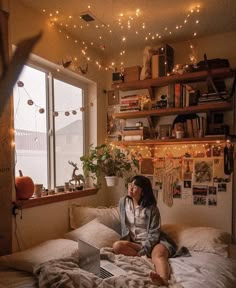 a woman is sitting on her bed with a laptop in front of her and lights hanging above her