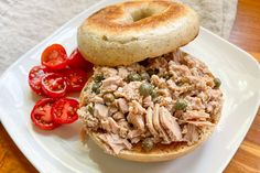 a sandwich with meat and vegetables on a white plate next to a bagel roll