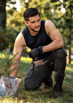 a man kneeling down next to a small tree in the grass with gloves on his hands