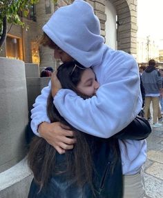 two people hugging each other in front of a building on a city street, one is wearing a hoodie and the other has her hair pulled back