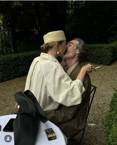 a man and woman are kissing while sitting on a chair in front of some bushes