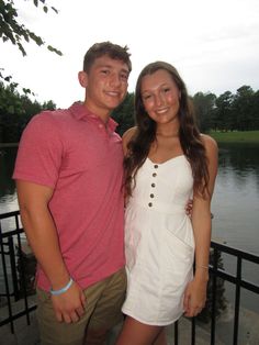 a young man and woman standing next to each other near a body of water with trees in the background