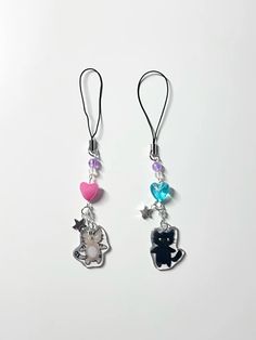 two earrings with charms attached to them on a white surface, one has a cat and the other has a heart