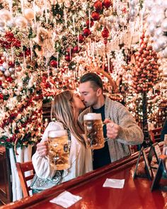 a man and woman kissing in front of a christmas tree holding mugs of beer