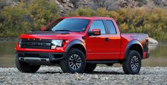 a red truck parked on top of a rocky beach next to a body of water