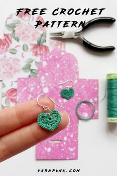 a hand holding a pair of green heart shaped earrings next to some thread and scissors