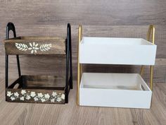 two wooden shelves with white flowers painted on the sides and gold handles, one shelf holding three small trays