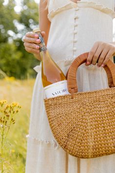 a woman holding a bottle of wine in her hand and a wicker basket on her shoulder