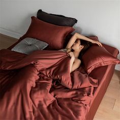 a woman laying on top of a bed covered in red sheets and pillows with her arms behind her head