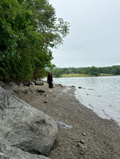 a person standing on the shore of a lake next to some rocks and trees with water in the background