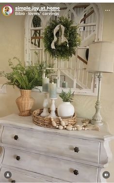 a white dresser topped with lots of greenery next to a clock and lamp on top of it