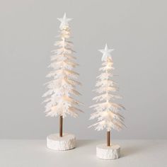 two small white christmas trees with lights on each one, standing in front of a gray background